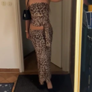 Leopardmönstrad sett - Säljer en snygg leopardmönstrad långklänning med omlott-detalj och knyt i midjan. Klänningen är axelbandslös och har en cool cut-out vid midjan. Perfekt för dig som vill sticka ut med ett djuriskt mönster och trendig passform.