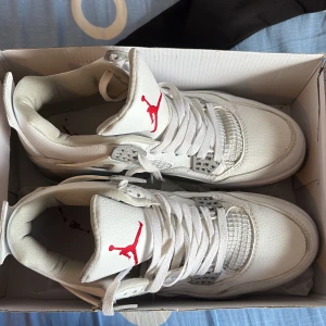 Nike Air Jordan 4 Retro White/Red - Säljer ett par Nike Air Jordan 4 Retro i vitt skinn med röda detaljer på tungan och innersulan. Skorna har klassisk meshpanel, gråa accenter och snörning. Perfekta för dig som vill sticka ut med en ikonisk sneaker med snygg design.