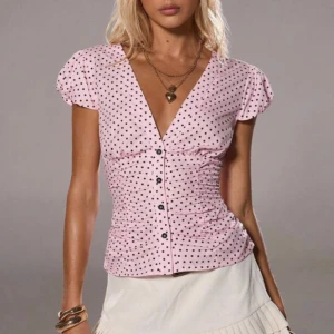 Rosa prickig blus med v-ringning - Supersöt rosa blus med svarta prickar och v-ringad hals. Korta puffärmar och knappar framtill ger en feminin vibe. Figurnära passform med lätt rynkade detaljer på sidorna. Perfekt för dig som gillar att sticka ut med färg och mönster.
