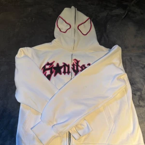 Vit Spider Gwen hoodie från Bongeer - Säljer en vit hoodie från Bongeer med Spider Gwen-inspirerad design. Tröjan har svart och rosa tryck med spindelmotiv på ryggen och coolt grafiskt tryck på bröstet. Huva med ögon-detaljer, dragkedja framtill och fickor. Perfekt för dig som gillar Marvel och streetwear.