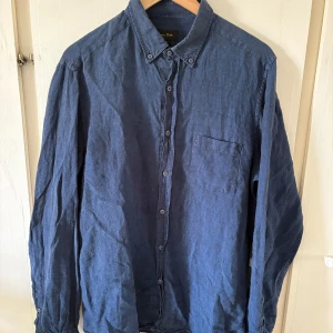 Blå linneskjorta från Massimo Dutti M - Snygg blå skjorta i 100% linne från Massimo Dutti. Klassisk modell med knappar hela vägen, bröstficka och krage med knappdetaljer. Perfekt för dig som gillar en avslappnad men stilren look.