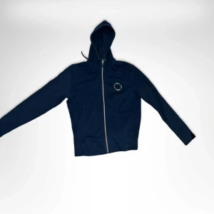 Mörkblå hoodie från Hugo boss  - Mörkblå hoodie från Hugo boss med dragkedja framtill och liten vit logga på bröstet. Tröjan har huva med snörning och långa ärmar. Perfekt för chill dagar eller när du vill ha en clean och sportig look.