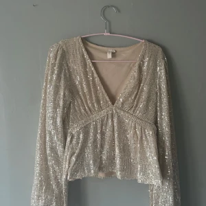 Glittrig champagnefärgad blus Nelly - Supersnygg glittrig blus från Nelly i champagnefärg med paljetter över hela toppen. Djup v-ringning, långa ärmar och knyt i midjan. Perfekt för dig som vill sticka ut med en festlig look. Croppad modell som ger en trendig siluett.