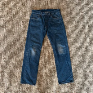 Levi's 501 blå jeans W32 L32 - Klassiska Levi's 501 jeans i blå denim med raka ben och normal passform. Byxorna har fem fickor, knappgylf och snygga slitningar på knäna och baksidan. Perfekta för en avslappnad och tidlös stil.