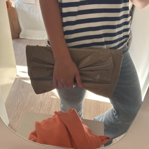 Beige kuvertväska med stor rosett - Säljer en beige kuvertväska i glansigt syntetmaterial med en stor dekorativ rosett på framsidan. Väskan har en stilren och modern look, perfekt att bära i handen. Enkel att matcha med olika outfits och rymmer det viktigaste du behöver.