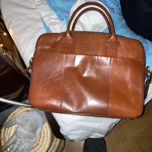 Brun datorväska i skinn från Bläck - Snygg brun datorväska från Bläck i äkta skinn. Väskan har två rejäla handtag, avtagbar axelrem och dragkedja upptill. Insidan har flera fack och detaljer i samma bruna ton. Perfekt för laptop och viktiga papper.