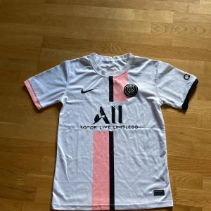 PSG tröja Nike xs/s - Snygg PSG fotbollströja från Nike i vit med rosa och svart vertikal rand på framsidan. Kortärmad med rund hals och svarta detaljer på ena ärmslutet. Materialet är lätt och andas, perfekt till vardags eller träning. Paris Saint-Germain-logga på bröstet och sponsortryck.