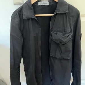 Svart Stone Island Overshirt - Svart Stone Island overshirt med dragkedja och knappar.  Jackan har en ficka på bröstet och Stone Island-märket på vänster ärm. Storlek: Small Nypris 3500kr I mycket fint skick.