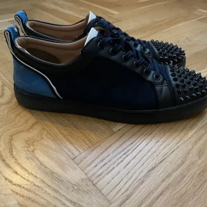 Mörkblå  christian louboutin med nitar - Snygga mörkblå sneakers från Louboutin med svarta nitar på tån, svarta skinn- och mockadetaljer samt klassisk röd sula. Skorna har snörning och cool kontrast med vit kant på hälen. Perfekta för dig som vill sticka ut. Priset kan diskuteras vid snabb affär.