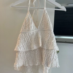 Vit volangig linnetopp med spets - Supersöt vit linnetopp med volanger och tunna axelband som knyts i nacken. Toppen har ett vågigt spetsmönster och är ärmlös med v-ringning. Perfekt för varma dagar när du vill ha en luftig och romantisk look. Bra skick!