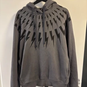 Grå hoodie Neil Barrett med blixtar - Snygg grå hoodie från Neil Barrett med ikoniskt svart blixtmönster över axlar och bröst. Tröjan är i nyskick! Nypris 6 793kr på Farfetch.  