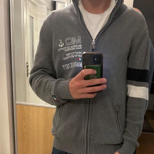 Grå kofta från Marina dept - Säljer en grå kofta med dragkedja från Marina dept. Tröjan har vita och svarta detaljer på ena ärmen, broderad text och ankare på bröstet samt ribbade muddar. Cardigan med Fickor med dragkedja och hög krage ger en chill och snygg look.