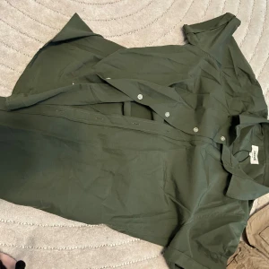 Mörkgrön kortärmad skjorta från zalando - Snygg mörkgrön skjorta från zalando med korta ärmar och klassisk krage. Skjortan har knäppning framtill med diskreta knappar och är gjord i ett lätt bomullstyg. Perfekt för en clean och avslappnad look.  Kartongen är kvar