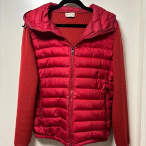 Röd Moncler cardigan med huva - Snygg röd pufferjacka från Moncler med stickade ärmar och quiltad front. Jackan har dragkedja, huva och två fickor framtill. Perfekt för dig som vill ha en sportig och trendig look med premiumkänsla.