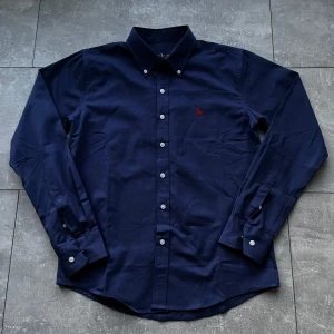 Ralph Lauren Skjorta - Klassisk mörkblå skjorta från Ralph Lauren med button-down krage och vit knappstängning. Perfekt för en clean och stilren look.