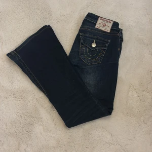 True Religion Joey Low Rise Flare Jeans - Mörkblå jeans från True Religion, modell Joey Low Rise Flare. Snygga kontrastsömmar, utsvängda ben och coola fickdetaljer med knappar bak. Klassisk femficksdesign och låg midja. Perfekta för dig som gillar retrovibbar och statement-jeans.