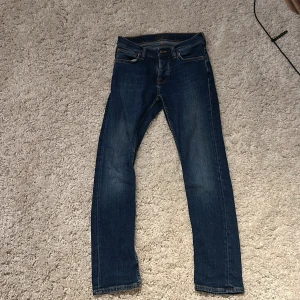 Mörkblå jeans från Nudie Jeans Co - Säljer ett par mörkblå jeans från Nudie Jeans Co i klassisk femficksmodell. Jeansen har snygga kontrastsömmar och broderade bakfickor med Nudie’s signaturvåg. Modellen är slim och materialet är mjukt denim i bomull.