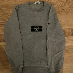 Grå sweatshirt från Stone Island - Säljer en grå långärmad sweatshirt från Stone Island med den klassiska logotypen på bröstet. Tröjan har rund hals och mjukt material som är skönt mot huden. Perfekt för dig som gillar streetwear och vill ha en clean look.