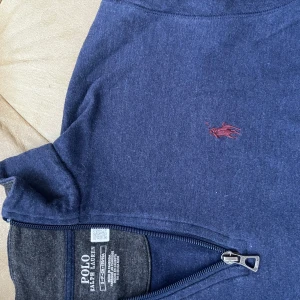 Blå half zip tröja från Polo Ralph Lauren - Snygg mörkblå långärmad tröja från Polo Ralph Lauren med half zip och klassisk röd broderad logga på bröstet. Tröjan har en mjuk bomullskänsla och ribbade muddar vid ärmslut och nederkant. Perfekt för chill dagar och enkel att matcha.