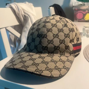 Gucci beige keps med GG-mönster - Säljer en beige keps från Gucci med klassiskt GG-monogram över hela kepsen. På sidan finns en snygg detalj med röd och marinblå rand. Justerbar rem bak i skinn och insida i mörkblått tyg. Perfekt för dig som vill ha en lyxig och trendig accessoar.