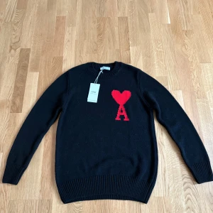 Svart stickad tröja från Ami Paris - Svart stickad tröja från Ami Paris med röd hjärtlogga och bokstaven A på bröstet. Klassisk rund halsringning och ribbade muddar. Perfekt för dig som gillar stilrena plagg med en twist. Materialet är mjukt och håller värmen under kyliga dagar.