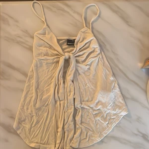 Vit linnetopp med knyt från Gina Tricot - Supersöt vit linnetopp från Gina Tricot med tunna axelband och snygg knytning framtill. Toppen har en lätt utsvängd passform och är perfekt för varma dagar. Enkel att styla med jeans eller kjol för en fräsch look.