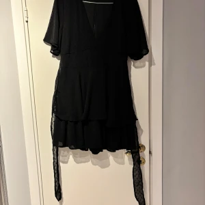 Svart kortklänning med volang - Svart kortklänning med v-ringning och volang nertill. Klänningen har korta ärmar och knytband i midjan. Inbyggda shorts.