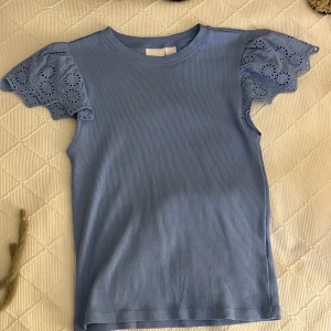 Blå topp med spetsärmar från Gina Tricot - Supersöt ljusblå topp från Gina Tricot med korta, volangiga spetsärmar. Toppen är ribbad i kroppen och har rund halsringning. Perfekt för en sommarlook🩷