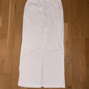 Vit långkjol med snörning och slits - Säljer en vit långkjol med diskreta vertikala ränder. Kjolen har snörning i midjan för att justera passformen och en snygg slits baktill som ger extra rörelsefrihet. Materialet känns mjukt och lätt, perfekt för varmare dagar.