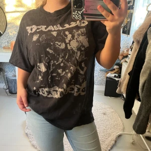 Oversized svart band-tshirt - Svart oversized t-shirt med coolt bandtryck framtill i grått och vitt. T-shirten har rund hals och korta ärmar, perfekt för dig som gillar streetstyle och vill ha en chill vibe. Materialet känns som mjuk bomull och passformen är loose.