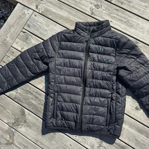Svart pufferjacka från Hangten 146/152 - Svart pufferjacka från Hangten med quiltad design och hög krage. Jackan har dragkedja framtill, två sidofickor med dragkedja och diskret logga på bröstet. 