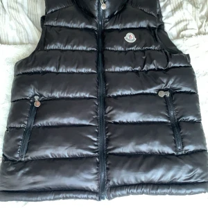Svart dunväst från Moncler - Snygg svart dunväst från Moncler med quiltad design och hög krage. Västen har två dragkedjefickor framtill och stängs med dragkedja. Perfekt att slänga över en hoodie eller tröja för extra värme. Klassisk Moncler-logga på bröstet, och den är oanvänd.