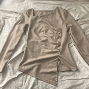 Beige långärmad omlottopp - Säljer en beige långärmad topp med asymmetrisk nederkant och snygg omlottdetalj framtill. Toppen har en stilren, figurnära passform och är tillverkad i ett mjukt, stretchigt material som känns skönt mot huden. Perfekt för dig som gillar minimalistisk och trendig stil.
