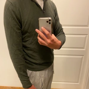 Olivgrön merinotröja half zip These Glory Days - Snygg olivgrön stickad tröja från These Glory Days i mjuk merinoull. Tröjan har half zip-dragkedja vid halsen och lång ärm, perfekt för lager-på-lager. Enkel och stilren design som funkar till det mesta.