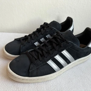 Adidas Campus sneakers svart/silver 42 2/3 - Snygga Adidas Campus sneakers i svart mocka med silvriga detaljer och klassiska tre ränder på sidorna. Skorna har vit sula, snörning och silvrig häl med Adidas-logga. Perfekta för dig som gillar en stilren och sportig look.