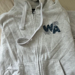 Ljusgrå hoodie/Russegenser - Ljusgrå hoodie med dragkedja och huva från Russeprofilering. Framsidan har blått WA-tryck med fyrverkerier och snörning i huvan. Baksidan har stort fyrverkeritryck och texten BOSVIK. Norsk Russegenser