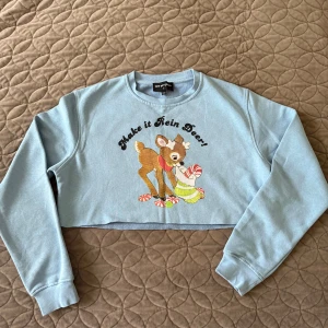 Croppad blå tröja med renmotiv (S) - Säljer en ljusblå croppad tröja från New Girl Order med gulligt renmotiv och texten 'Make it Rein Deer!' framtill. Tröjan har långa ärmar och rund hals. Materialet är mjuk bomull och polyester, perfekt för dig som gillar juliga prints och chill stil.