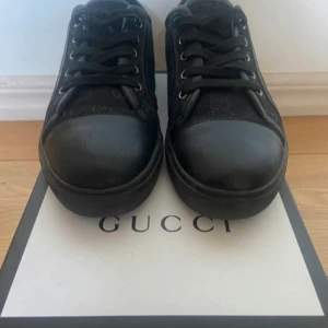 Svarta sneakers från Gucci - Stilrena svarta sneakers från Gucci med klassisk GG-monogram på sidorna. Skorna har rund tå, snörning och detaljer i skinn och textil. Perfekta för dig som vill ha en lyxig och clean look med diskret branding.