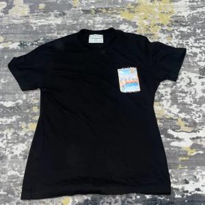 Svart Casablanca t-shirt med tryck - Svart t-shirt från Casablanca i bomull med färgstarkt grafiskt tryck av pool och sol på bröstet och ryggen. Trycket har inslag av orange, blått och vitt, och texten 'CASABLANCA' syns tydligt. Klassisk rund hals och korta ärmar. Perfekt för dig som gillar coola prints.