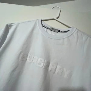 Vit Burberry t-shirt med logga - Snygg vit t-shirt från Burberry i bomull med klassisk rund hals och korta ärmar. Framsidan har en stilren, upphöjd Burberry-logga i vitt för en clean och exklusiv vibe. Perfekt för dig som gillar minimalistisk design och premiumkänsla. Använd 1 gång säljer för att den var för stor för mig.