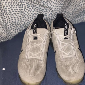 Nike Vapormax vita sneakers - Säljer ett par Nike Vapormax sneakers i vitt med stickad ovandel och tydlig svart swoosh på sidan. Skorna har genomskinlig sula med synliga luftbubblor och vita snören. Perfekta för dig som gillar sportig och modern stil.