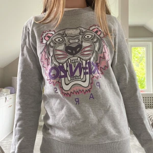 Grå Kenzo sweatshirt med tigertryck - Grå sweatshirt från Kenzo med ikoniskt broderat tigerhuvud i grått, rosa och lila på bröstet. Tröjan har rund hals, långa ärmar och ribbade muddar. Perfekt statement-piece för dig som gillar streetstyle och coola prints.