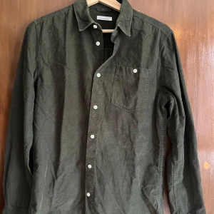 Grön manchesterskjorta Knowledge Cotton - Snygg grön manchesterskjorta från Knowledge Cotton Apparel. Skjortan har klassisk krage, vita knappar framtill och en bröstficka med knapp. Perfekt för dig som gillar en stilren och avslappnad look.