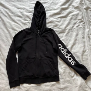 Svart Adidas hoodie med dragkedja - Svart hoodie från Adidas med stor vit logga längs ena ärmen. Tröjan har huva med snörning, dragkedja framtill och två fickor. Perfekt för chill eller sportiga dagar. Mjuk bomullskänsla och klassisk streetstil.