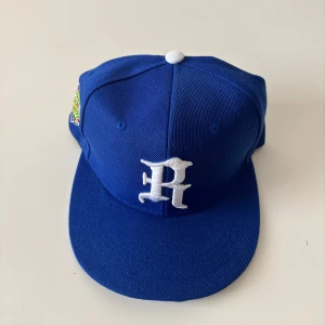 Blå baseball keps - Snygg blå keps med vit broderad bokstav på framsidan och färgglad brodyr på sidan. Kepsen har en klassisk snapback-modell med justerbar baksida och platt skärm. Perfekt för dig som vill sticka ut med en cool accessoar. Helt ny