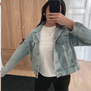 Ljusblå jeansjacka med snörningsdetaljer - Säljer en ljusblå jeansjacka med svarta snörningsdetaljer längs sidorna och slitna partier. Jackan har klassisk krage, knappar framtill och två bröstfickor. Perfekt för dig som vill ha en unik och trendig look.