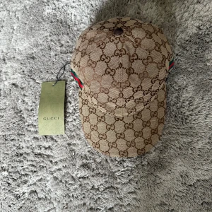 Gucci Keps - Exklusiv keps från Gucci i beige canvas med klassisk och lyxig design. En tidlös accessoar som kombinerar premiumkvalitet med stilren elegans – perfekt till både vardag och resa.  ♦️ Helt ny – med originaltag kvar ☑️ ⚜️ Unisex-modell – passar både herr & dam ☑️ ♦️ Ikonisk Gucci-design i beige canvas ☑️  📦 Snabb leverans & trygg affär 
