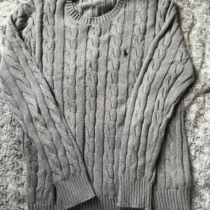 Grå kabelstickad tröja Polo Ralph Lauren - Snygg grå kabelstickad tröja från Polo Ralph Lauren med rund hals och långärmad passform. Tröjan har det klassiska broderade Polo-logot på bröstet och ribbade muddar vid ärmslut och nederkant. Perfekt för dig som gillar stilrena och tidlösa plagg.