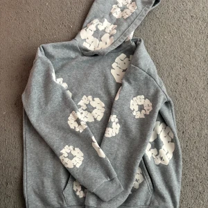 Denim tears  - Grå hoodie med vita blommiga tryck över hela tröjan. Tröjan har huva, långa ärmar och en klassisk känguruficka framtill. Mjuk och skön, perfekt för chill dagar eller när du vill sticka ut lite extra.