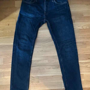 Mörkblå jeans från Nudie Jeans 30/32 - Säljer ett par mörkblå jeans från Nudie Jeans i klassisk femficksmodell. Jeansen har smal passform och raka ben (Grim Tim) med snygga kontrastsömmar och Nudie-logga på bakfickan. Perfekta för dig som gillar stilrena och tidlösa jeans.